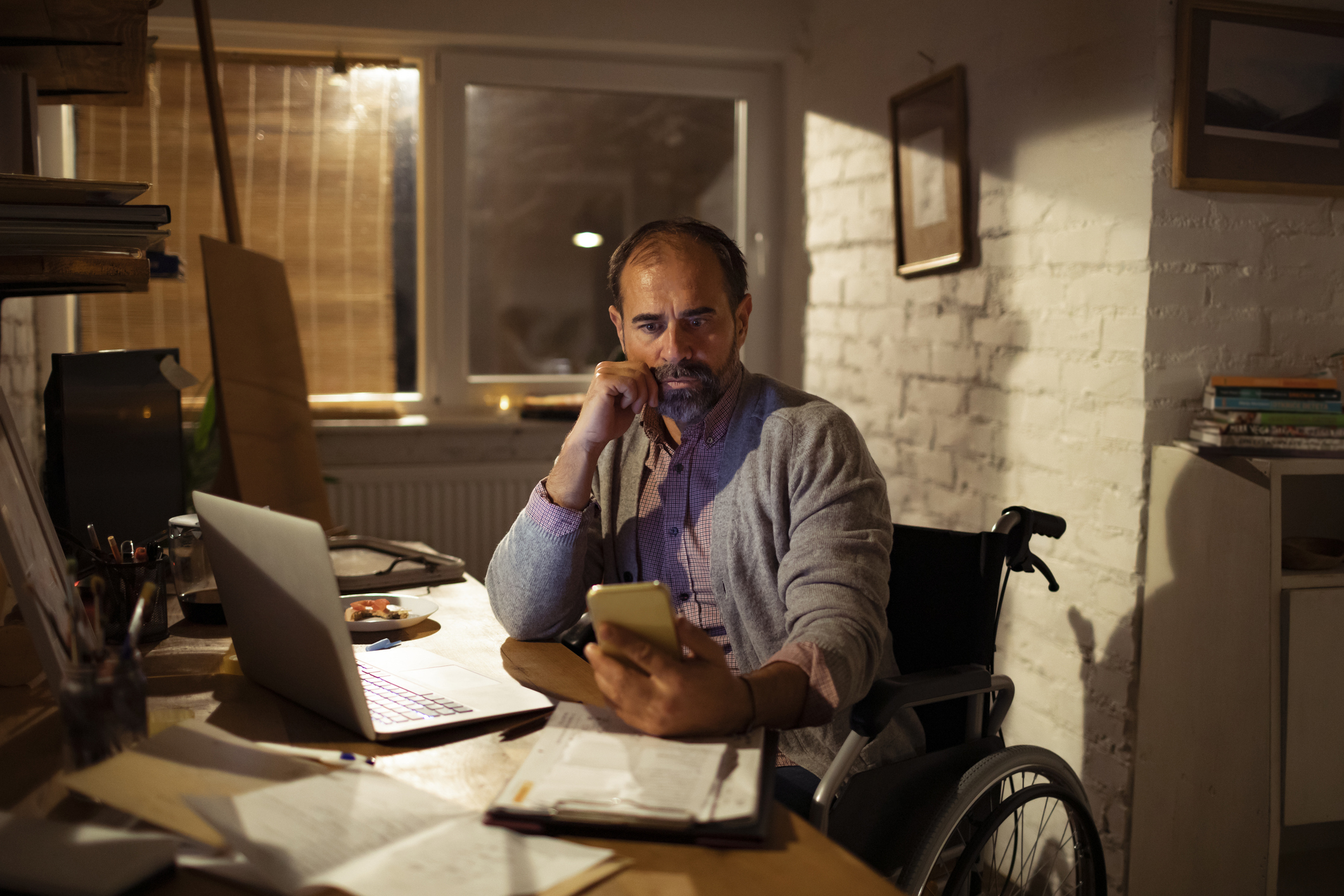 Man in wheelchair at home, working on paperwork with a calculator at his desk.