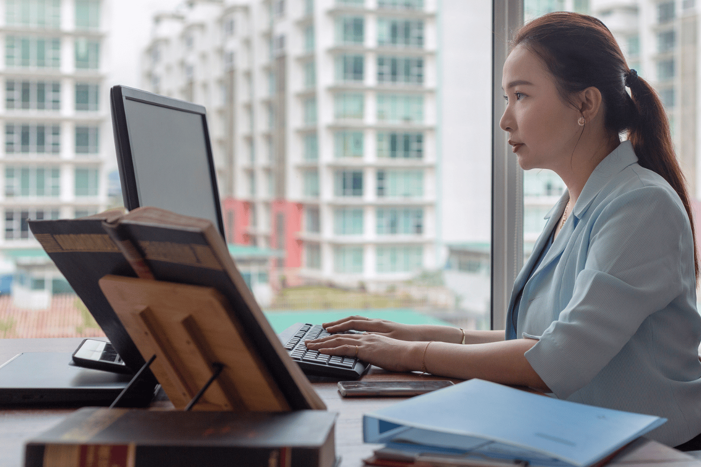 Woman in office working on computer with a city view in her background.