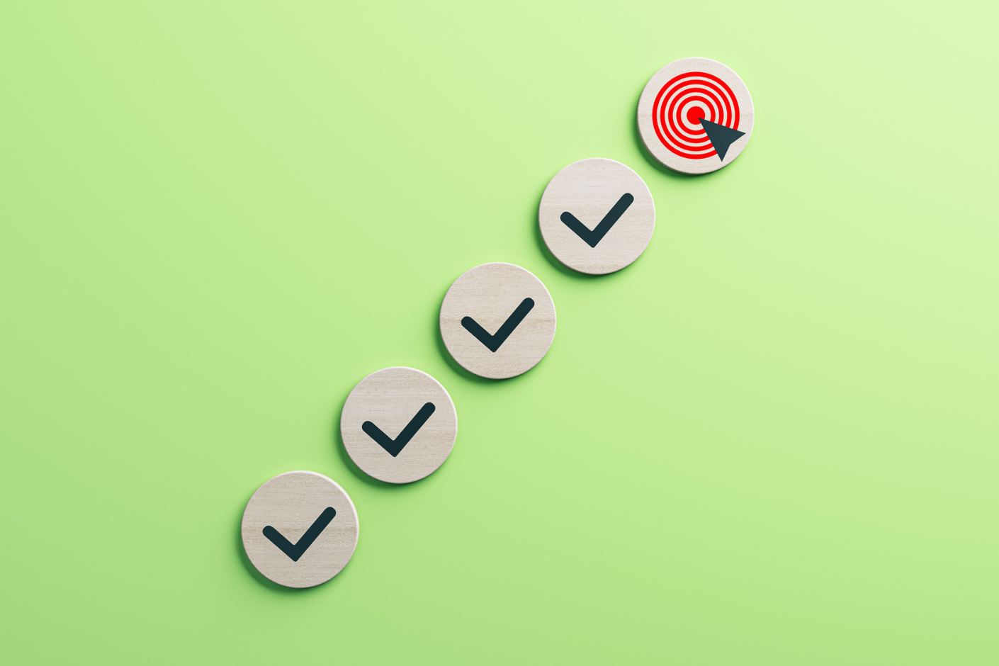 Circles of wood with black checkmarks laid out from left to right in a graduating, upward direction leading to a circle of wood with a red bullseye at the top right.