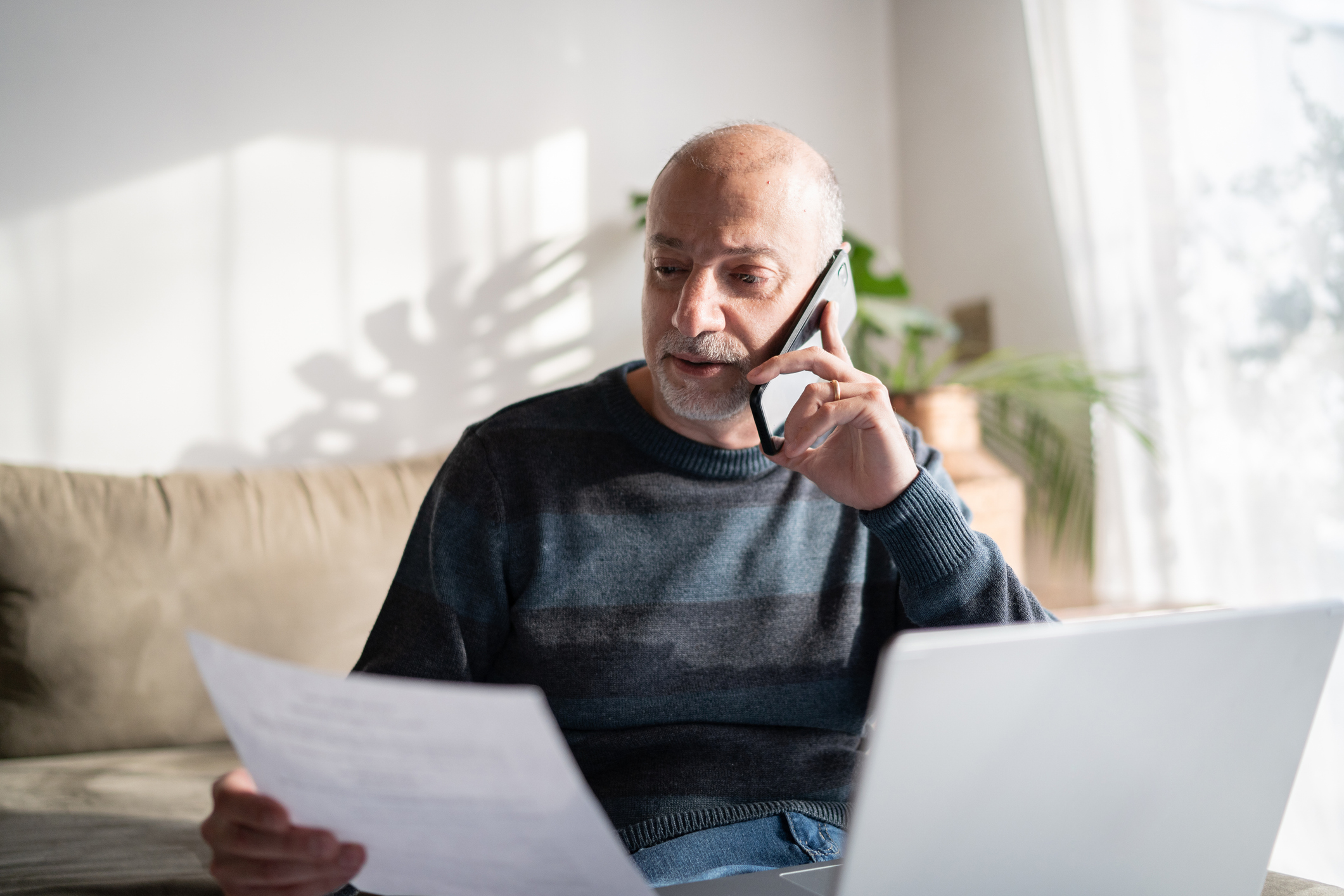 Middle-aged man in living room on couch, speaking on phone while looking at a document and at his laptop.