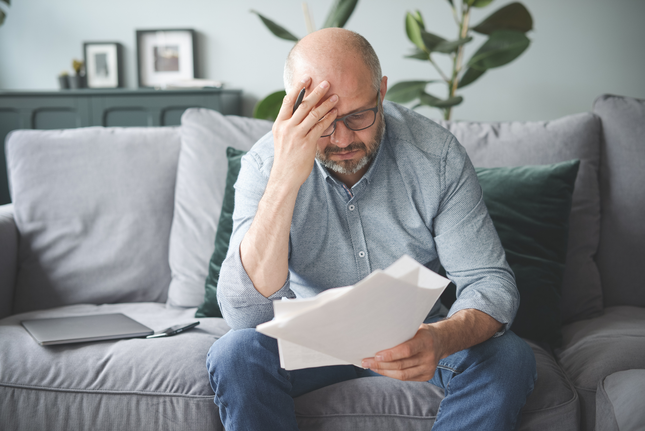Frustrated middle-aged man on his couch in his living room, looking at paperwork.