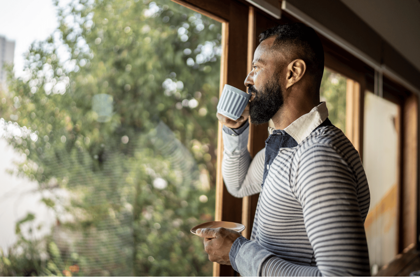 Man drinks coffee at home, standing at a window and looking outside.