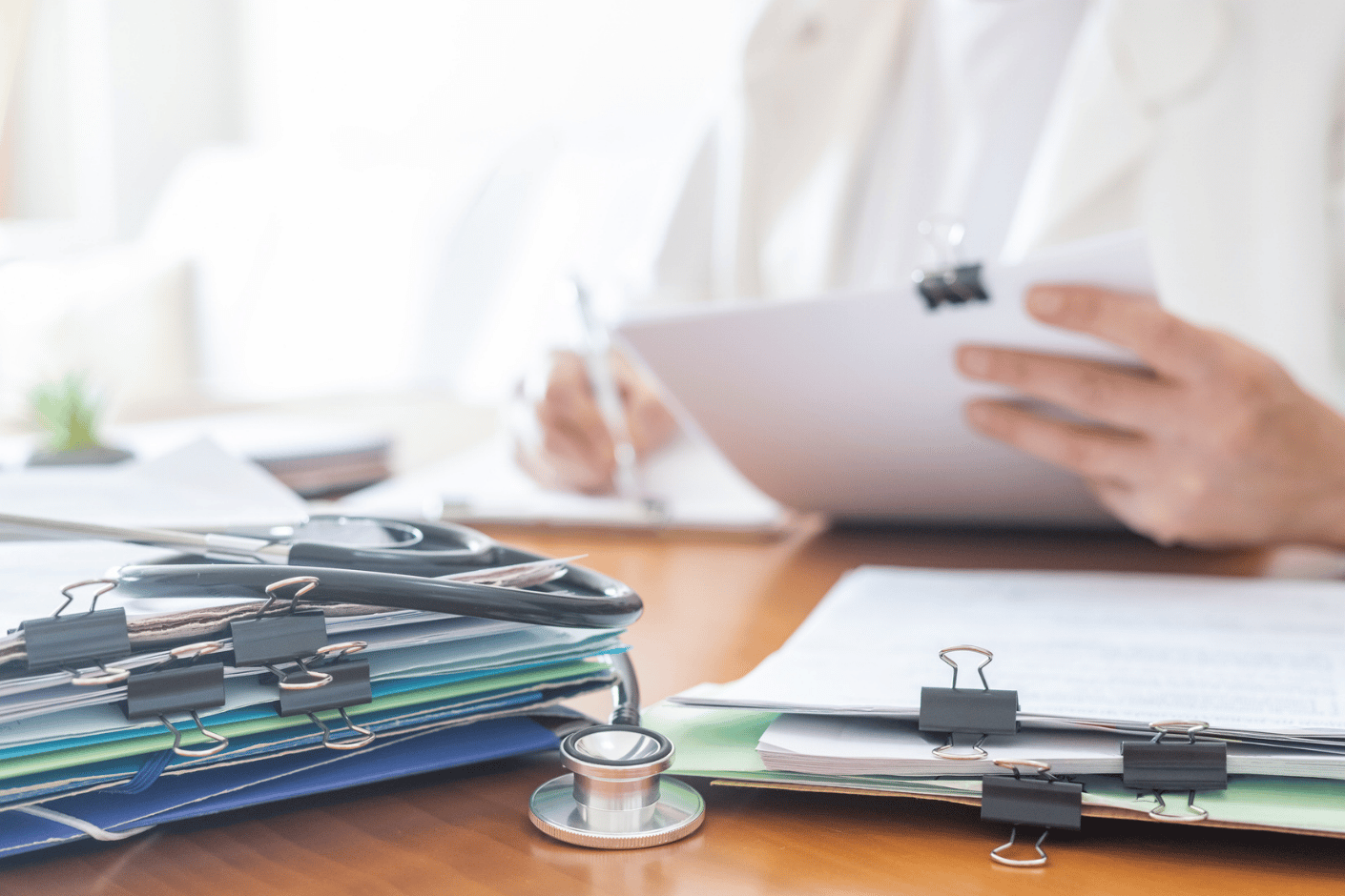 Doctor working at their desk with a pile of medical records, clipboard and a stethoscope in front of them.